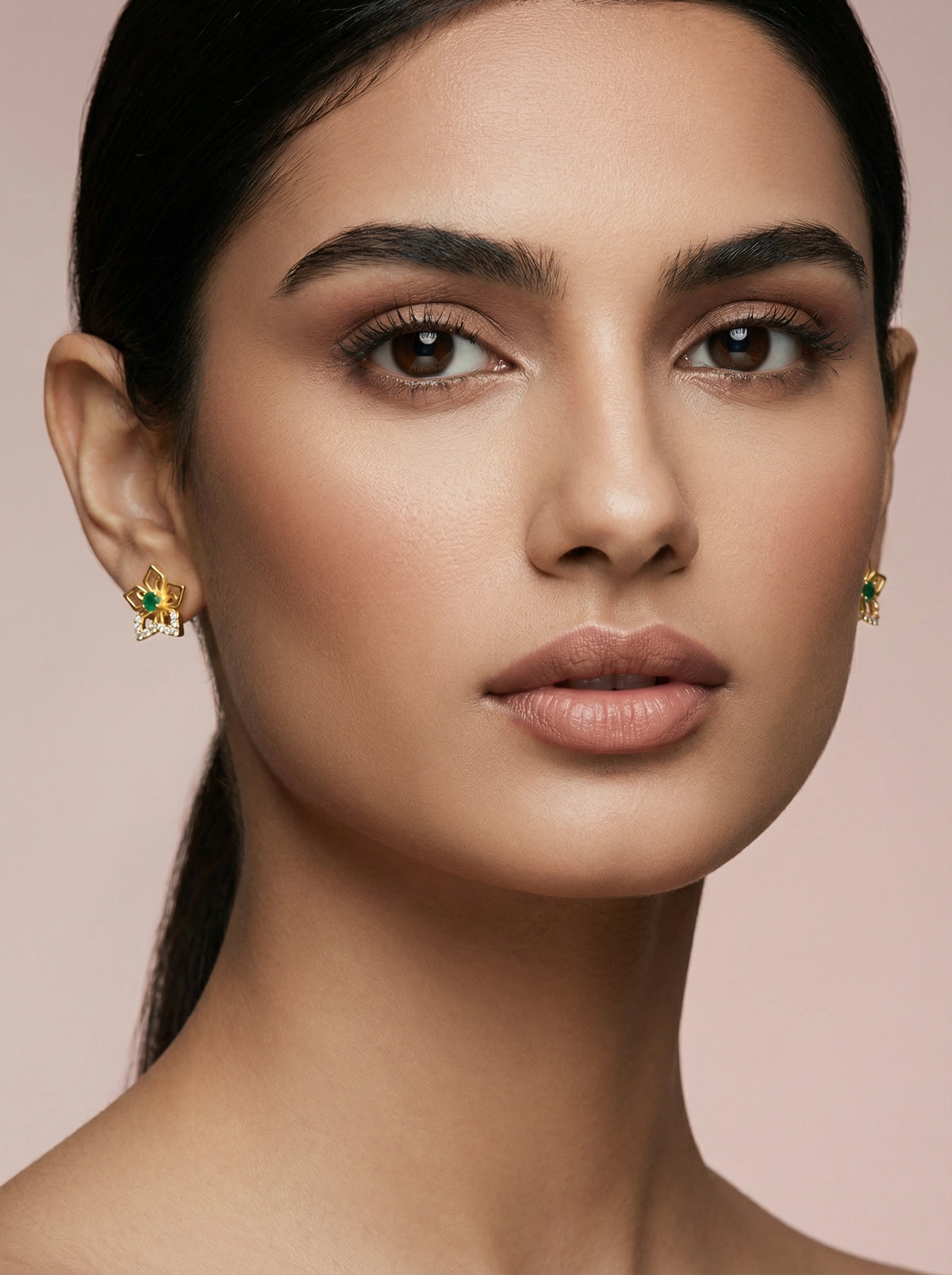 close-up of a woman wearing 18k  gold plated earrings with green gemstones on a pink background