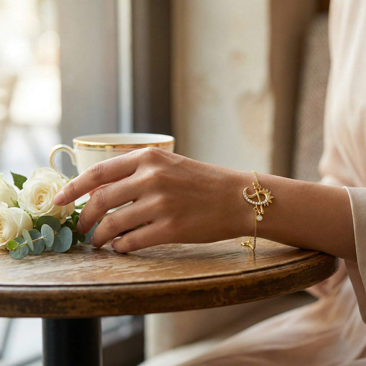 hand with 18k gold plated bracelet on a wooden table with flowers and a cup.