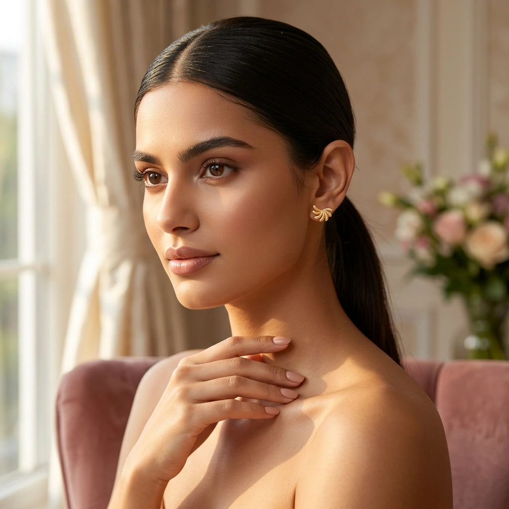 woman with a ponytail wearing 18k  gold plated earrings, sitting on a couch with a blurred floral arrangement in the background.