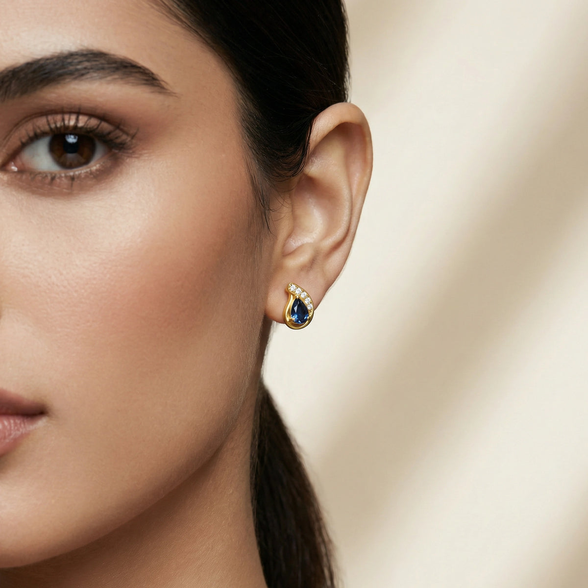 close-up of a woman wearing a 18k gold plated earring with a blue gemstone against a neutral background