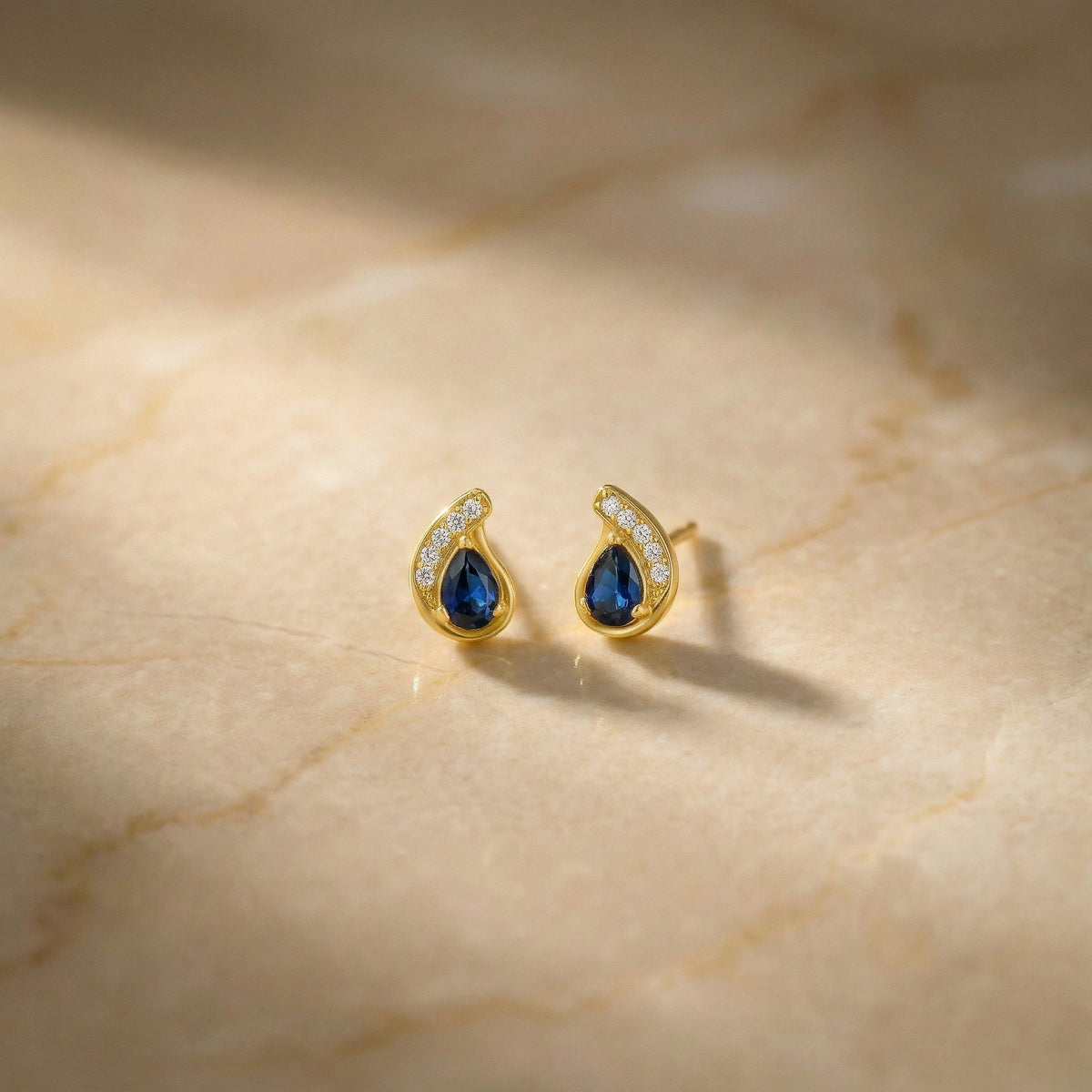 18k gold plated earrings with blue gemstones on a beige marble surface