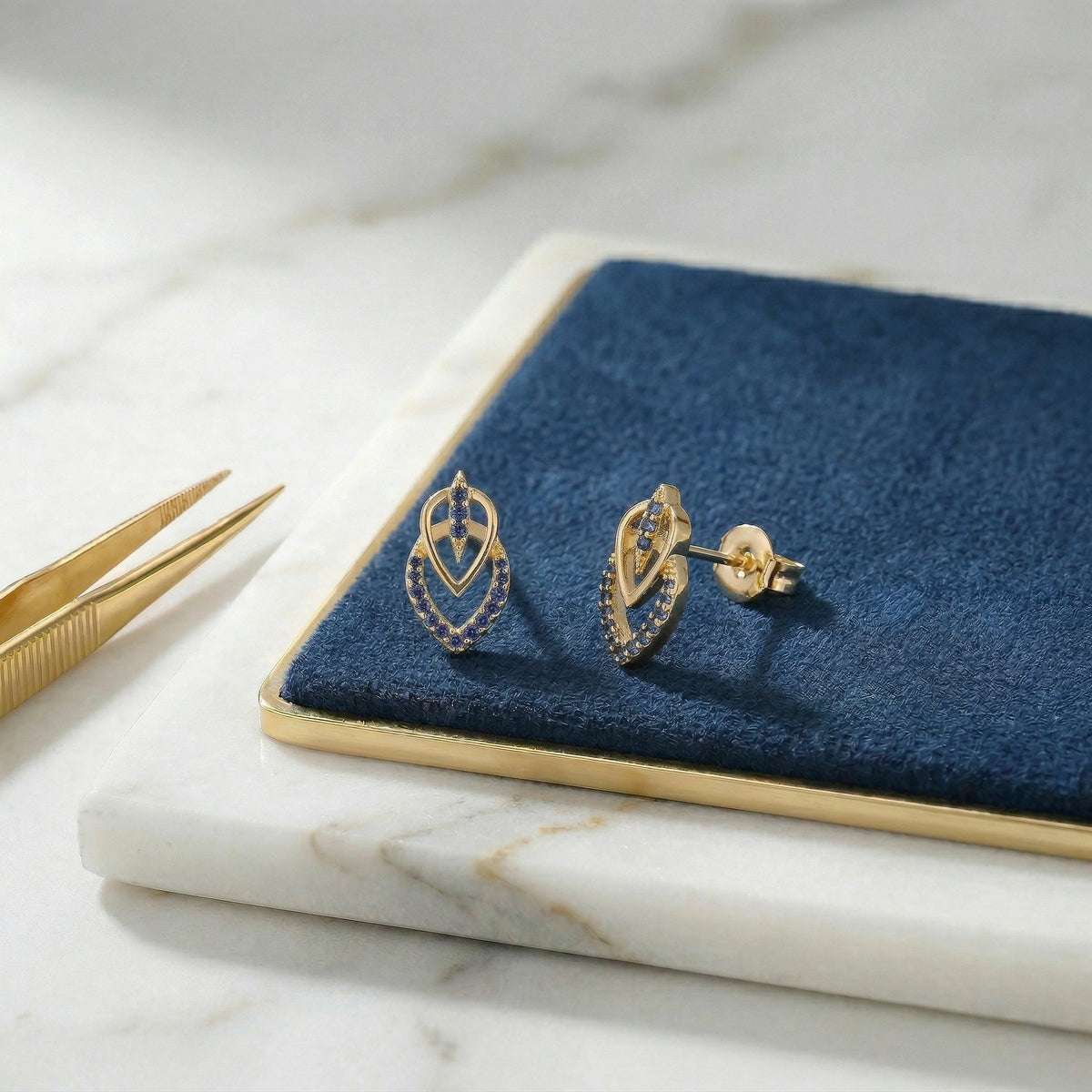 18k gold plated earrings on a blue velvet cushion with tweezers on a marble surface
