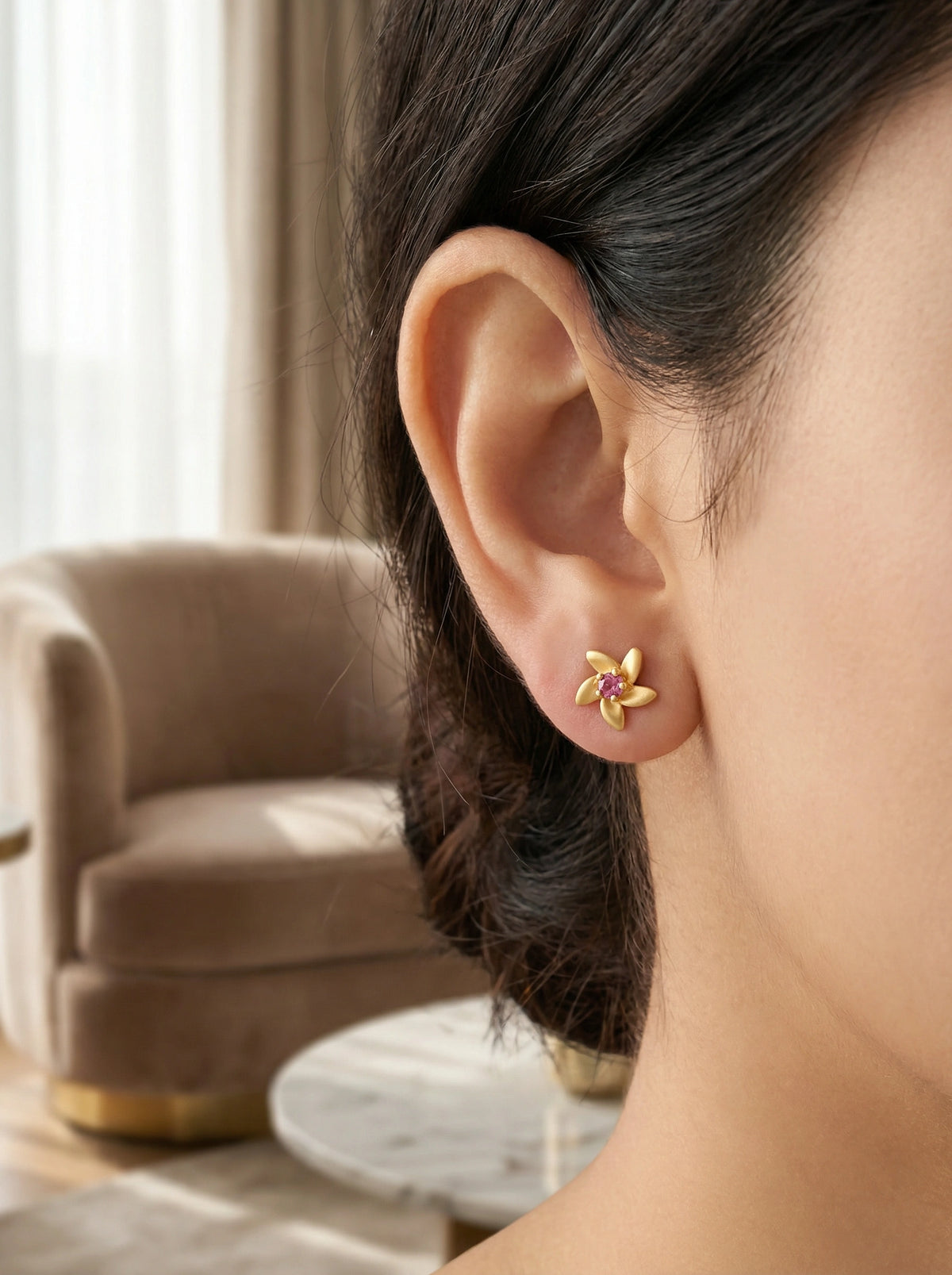 close-up of a person wearing 18k  gold plated floral earrings with a blurred indoor background.