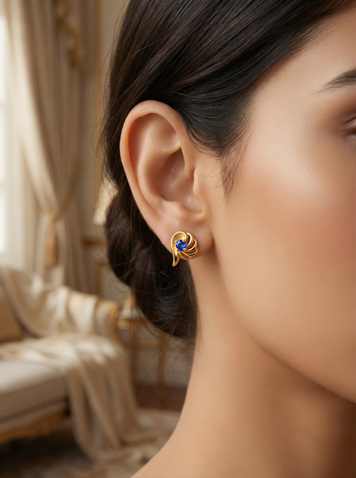 close-up of a woman wearing a 18k gold plated earring with a blue gemstone, blurred background