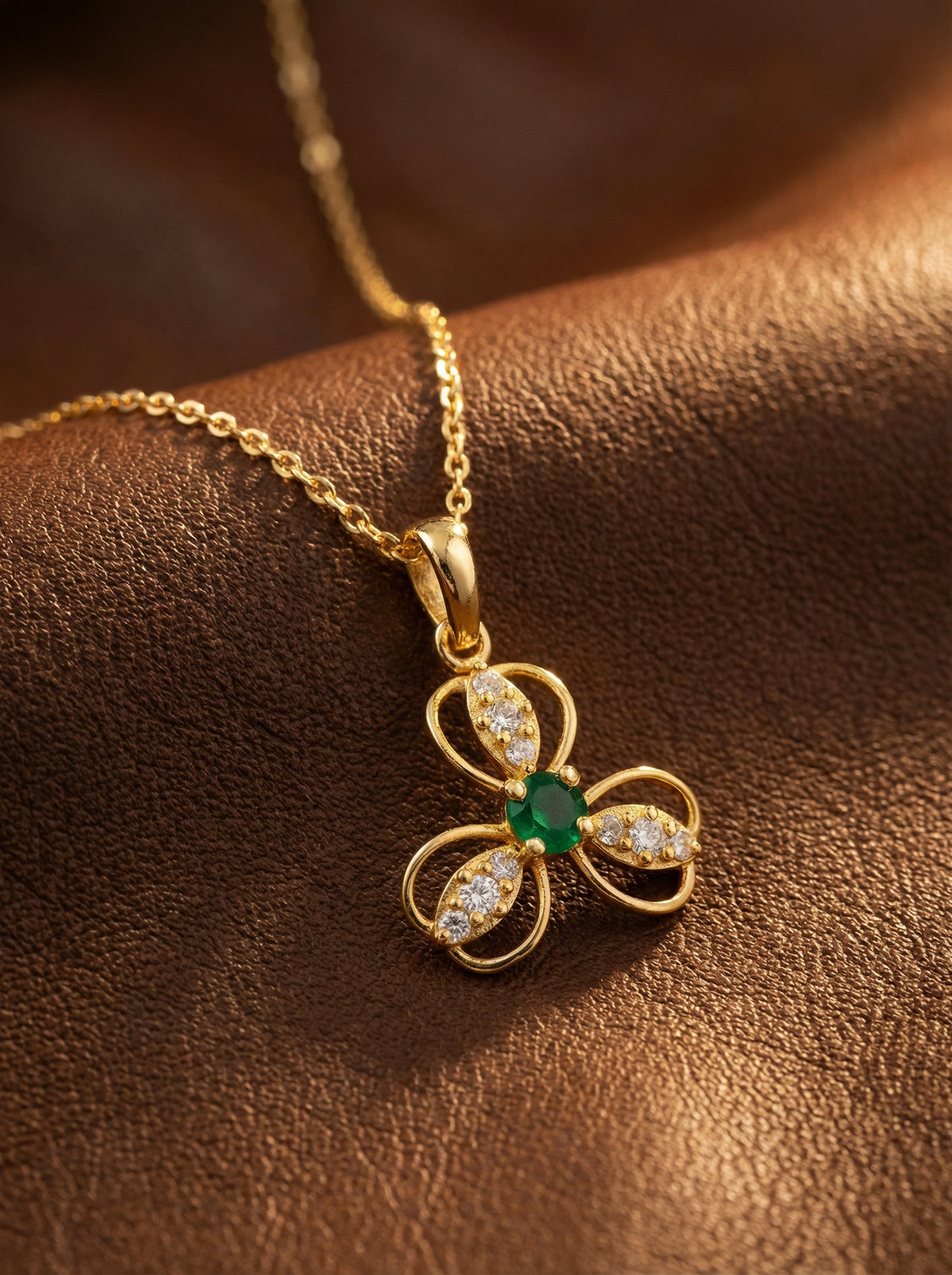18k gold plated necklace with a clover-shaped pendant featuring an emerald and diamonds on a brown leather surface.