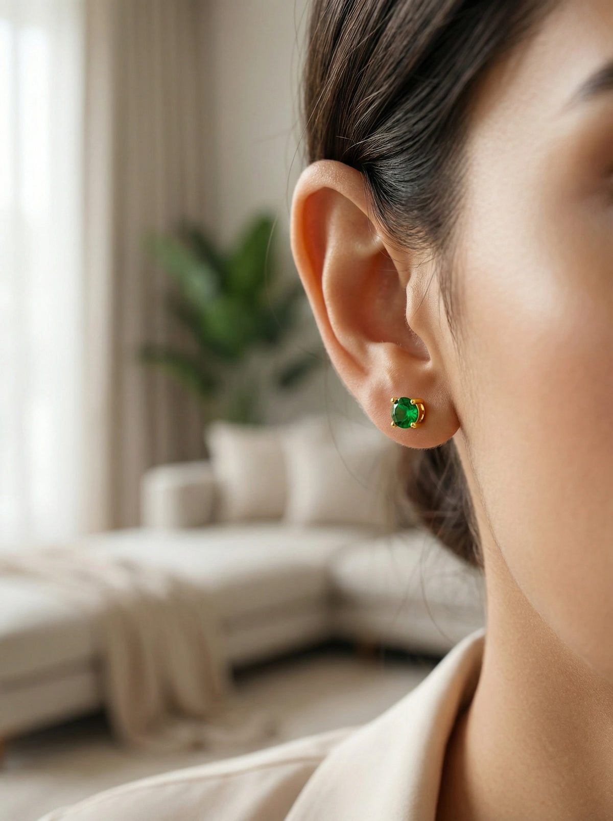 close-up of a person wearing green 18k gold plated earrings with a blurred indoor background