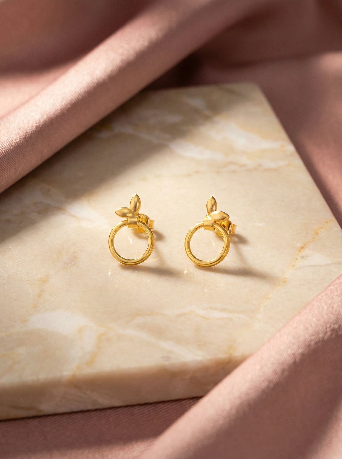 18k gold plated earrings on a marble surface with a soft pink fabric background