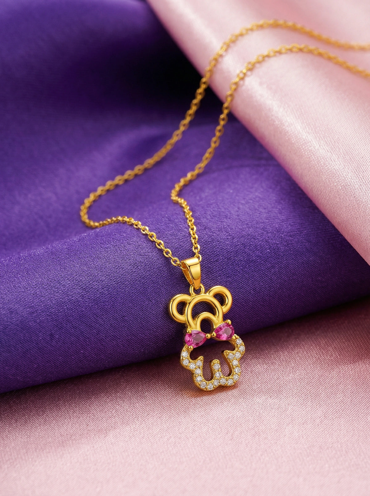 18k gold plated necklace with a teddy bear pendant featuring pink and purple stones on a purple and pink fabric background.