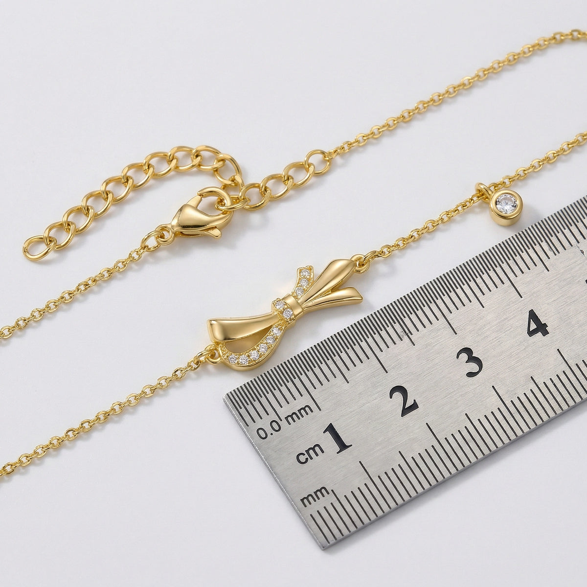 18k gold plated bracelet with a bow charm next to a ruler for size reference on a white background