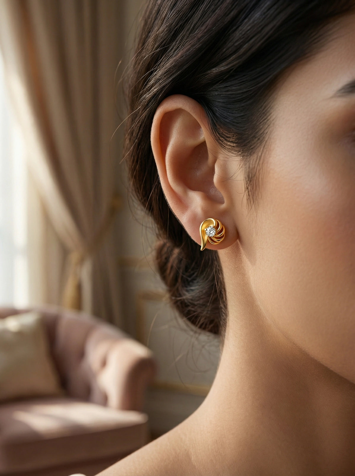close-up of a person wearing 18k gold plated  earrings with a diamond, set against a blurred indoor background.